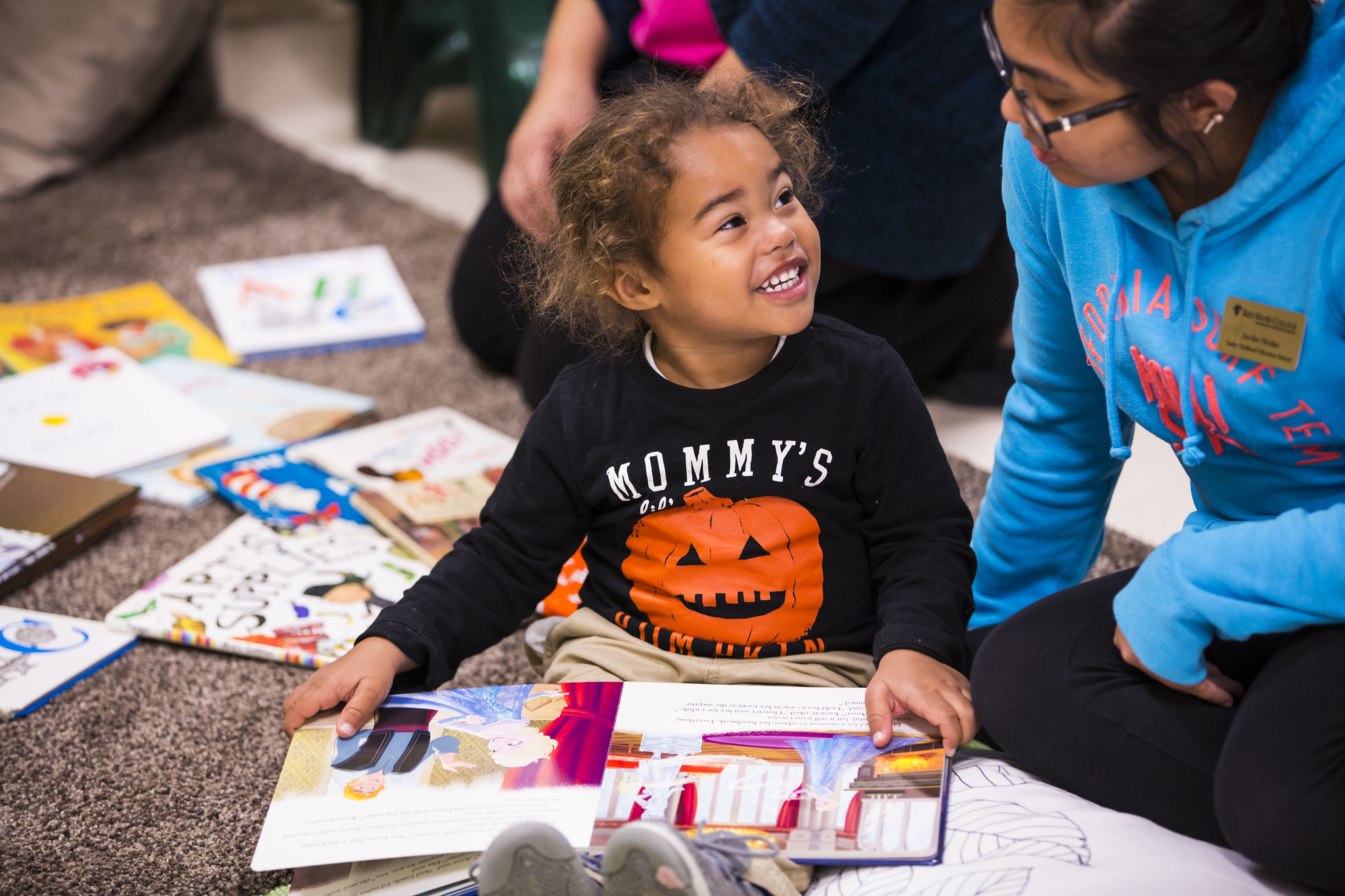 Young girl with pumpkin on shirt, smiling and surrounded by books in day-care space.