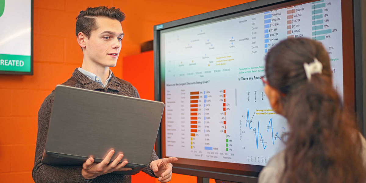 Students speak in front of a screen displaying charts and statistics.